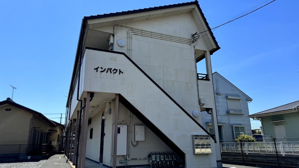 白い外壁の 2 階建ての建物の外観。手前に駐車場がある