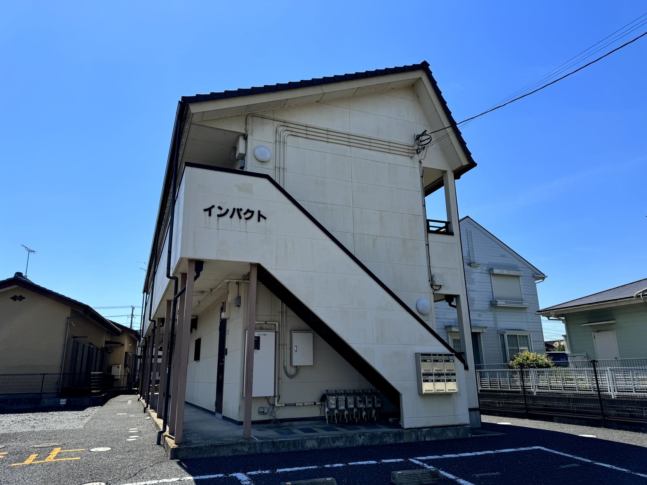 白い外壁の 2 階建ての建物の外観。手前に駐車場がある